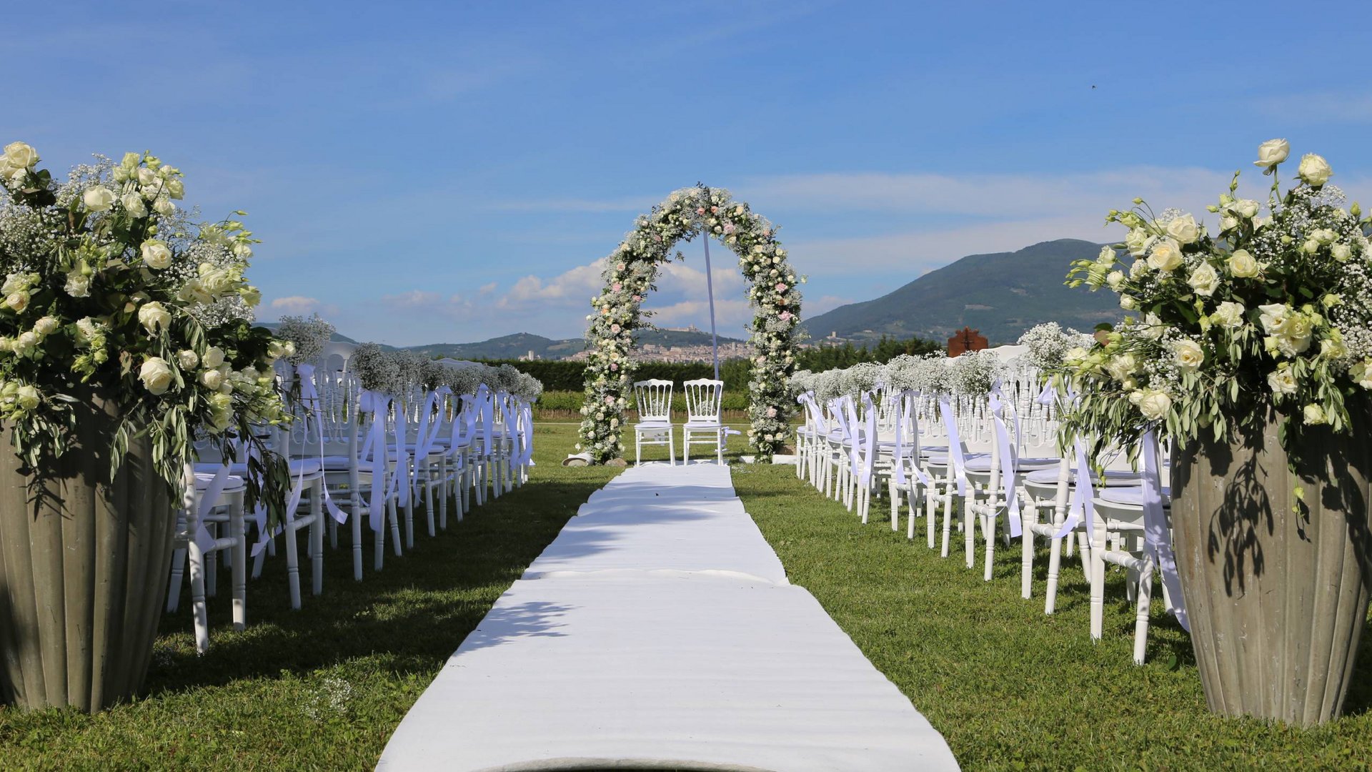 Valle di Assisi: un resort per matrimoni ad Assisi Allestimento esterno per cerimonia di matrimonio con sedie bianche e arco floreale