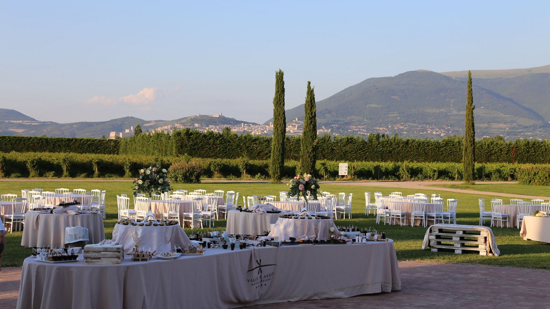 Valle di Assisi: un resort per matrimoni ad Assisi Tavoli all'aperto apparecchiati per un evento in giardino con colline sullo sfondo
