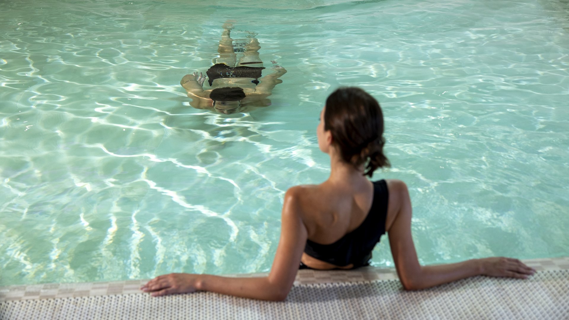 Assisi Cantico SPA - La tua SPA ad Assisi, Perugia, nell' Umbria più bella! Percorsi Benessere Massaggi Idee Regalo per Adulti e Bambini Donna in costume da bagno nera si rilassa a bordo piscina con altra persona che nuota