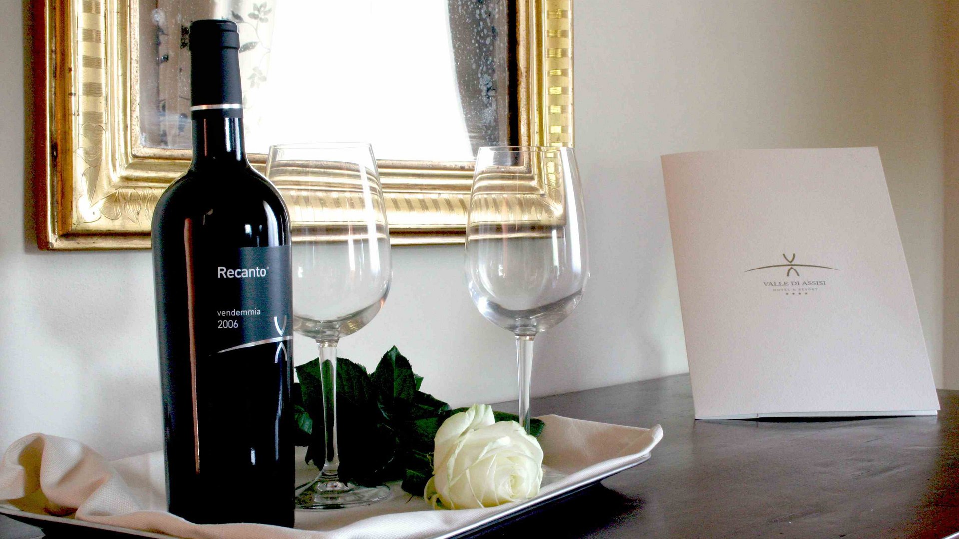 Hotel a 10 minuti di macchina da Assisi e la Basilica di San Francesco e il suo centro storico, 3 minuti di macchina dalla Porziuncola Bottiglia di vino rosso con due calici e rosa bianca su vassoio