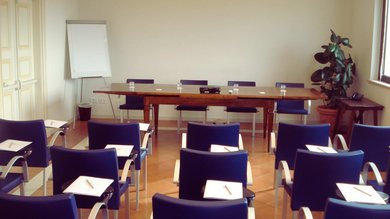 A Santa Maria degli Angeli - Assisi ( Perugia ) Centro Congressi e Meeting d'eccezione? I tuoi eventi vista Assisi Umbria Aula conferenze con sedie blu e tavolo con bicchieri e lavagna a fogli mobili