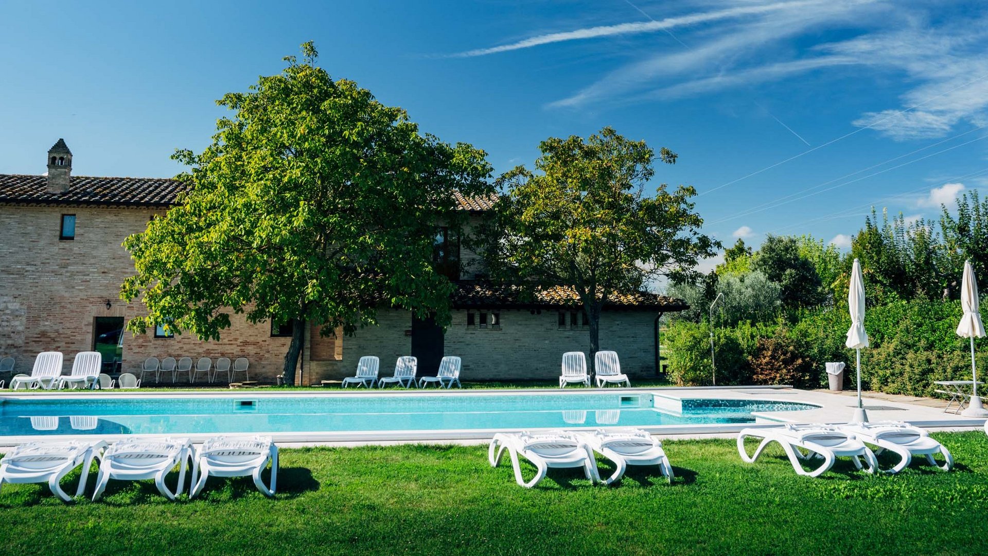 Ferienhaus in Umbrien mit Pool: in unserem Resort Außenpool mit Liegestühlen und Sonnenschirmen in einem sonnigen Garten vor einem Haus