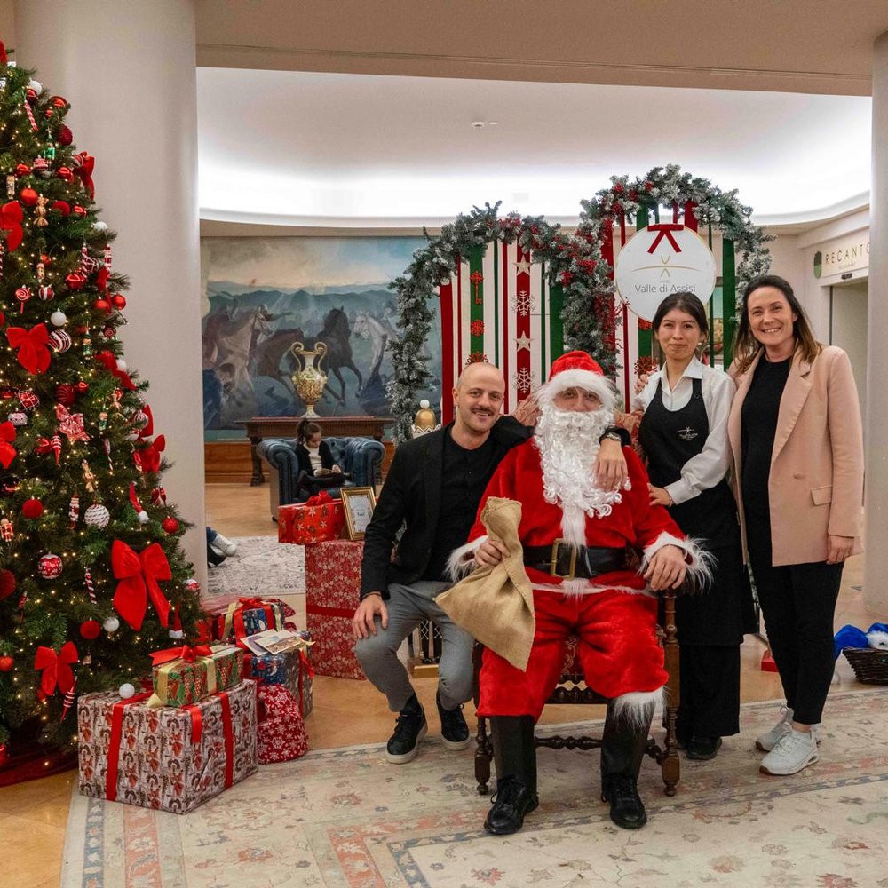 Natale e Inizio Anno a Valle di Assisi 2025 Persone sorridenti con Babbo Natale accanto a un albero di Natale decorato e regali